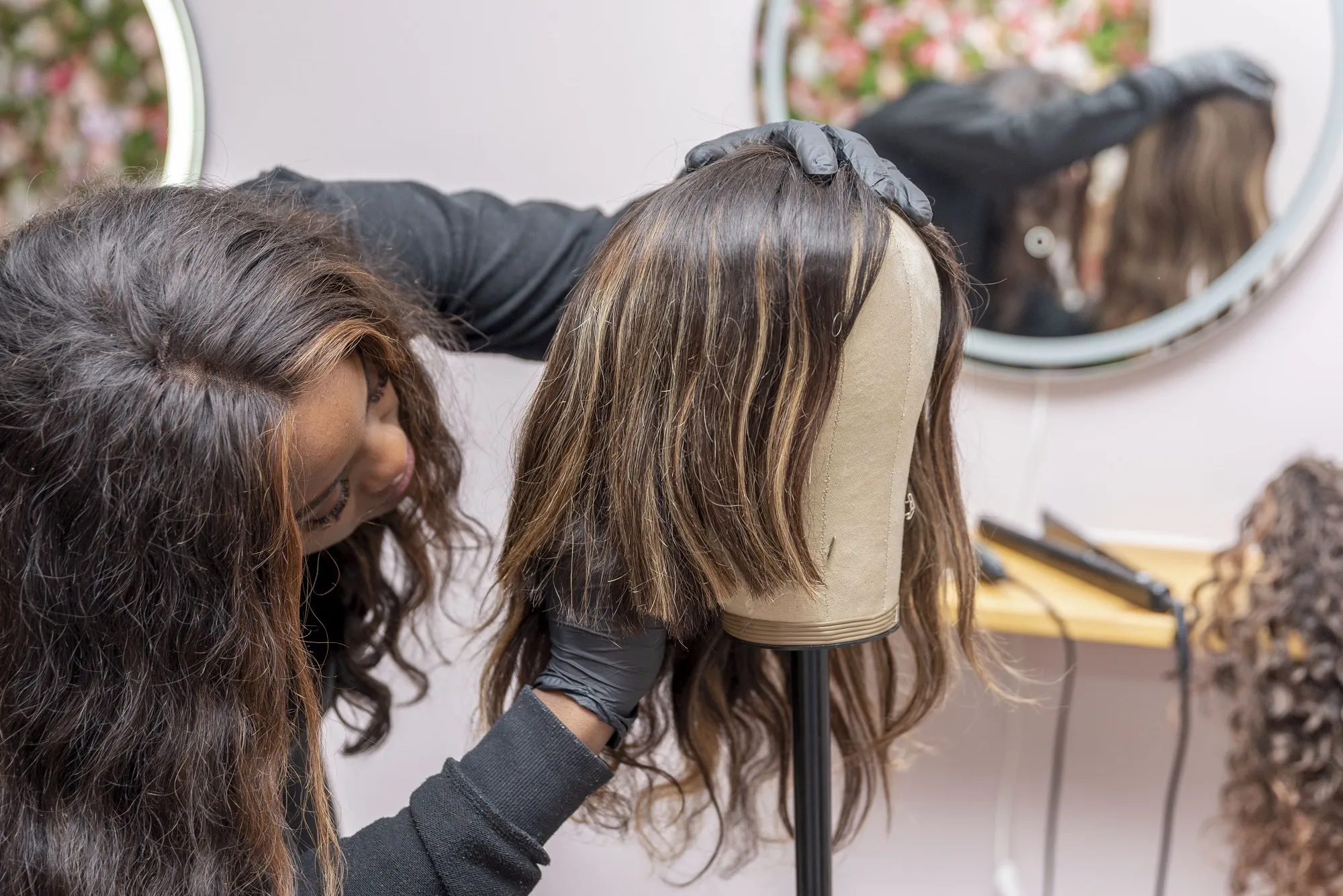 Specialist met zwarte handschoenen werkt aan een lang, bruin haarwerk met karamelkleurige highlights op een paspop.